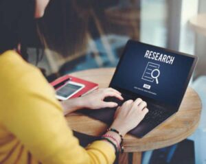 A woman in a yellow sweater types on a laptop that shows a screen that says "research"